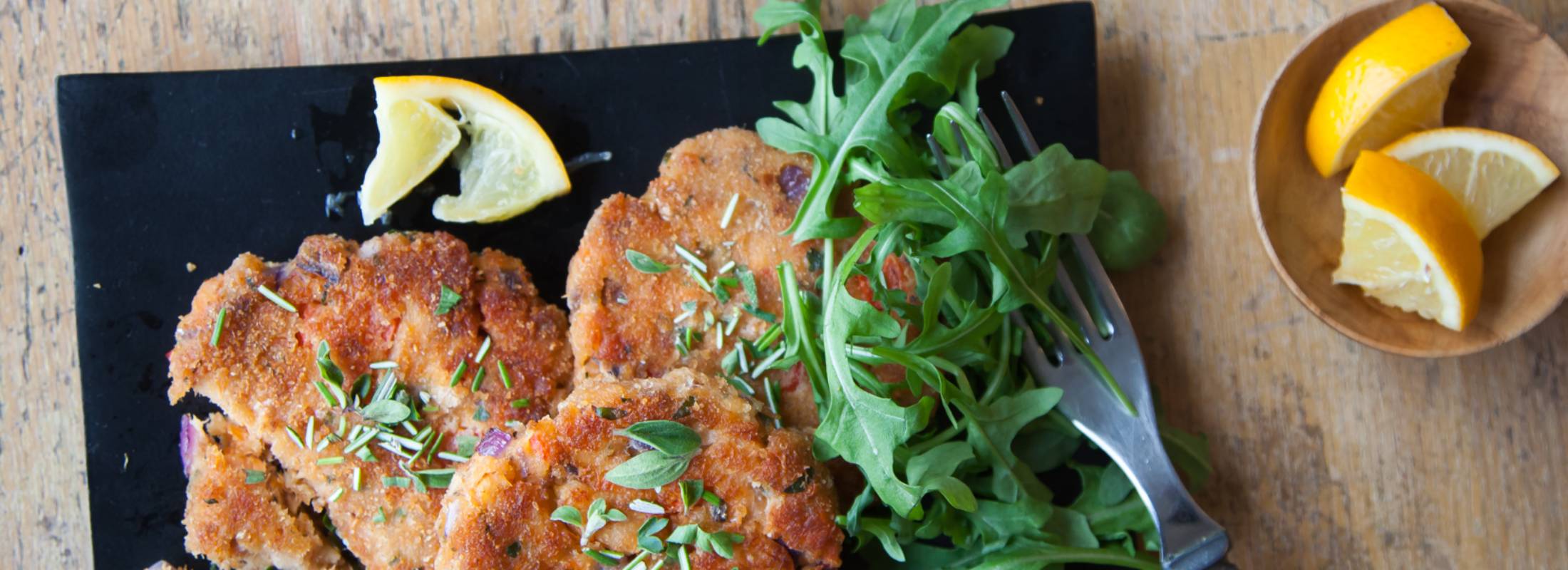 Wild Salmon Cakes on a black plate with arugula, old fork next to a bowl of lemon wedges
